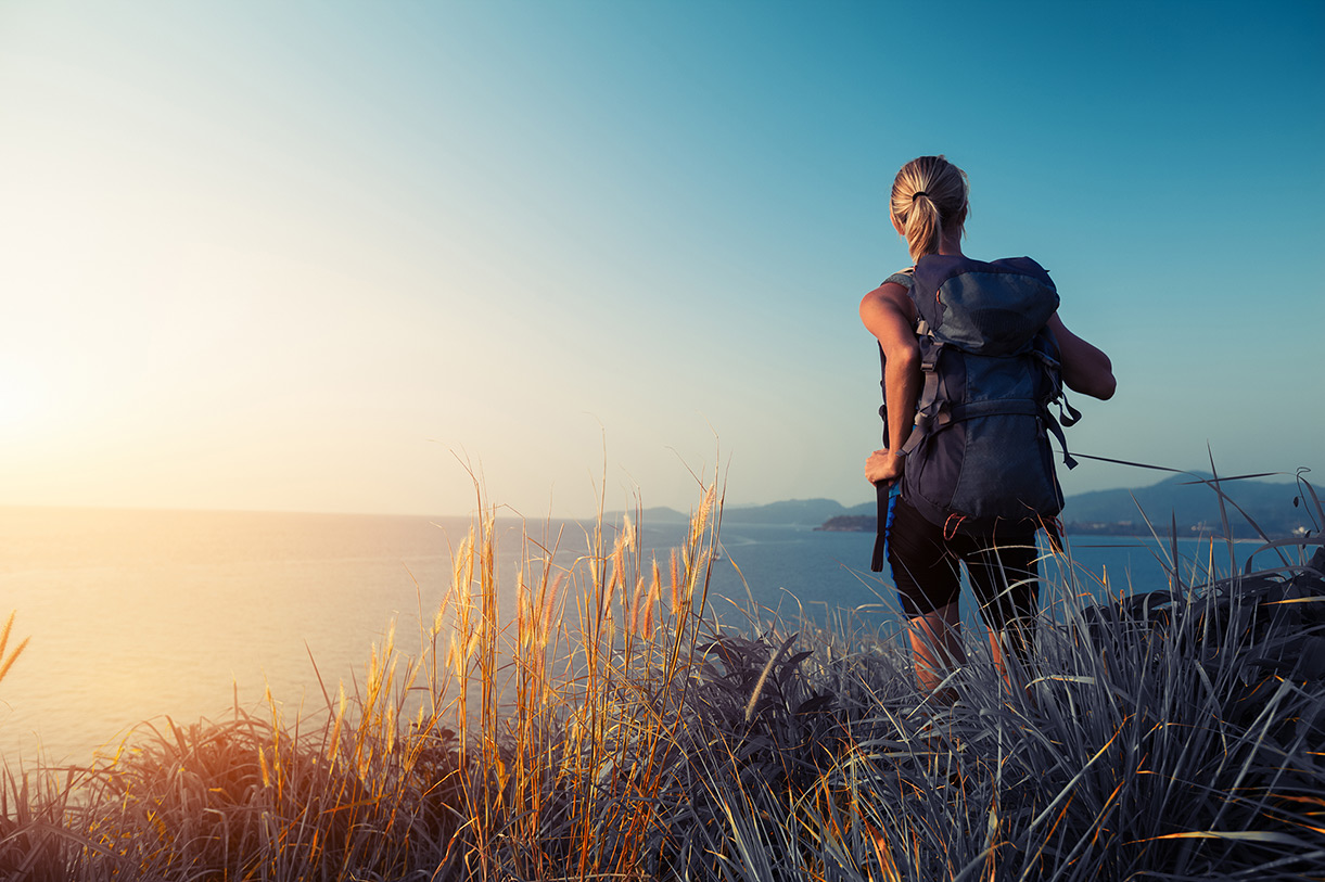 Woman with backpack hiking by ocean and mountains on a sunny day